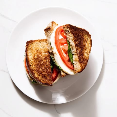 Fresh basil and juicy tomato slices peek from the crispy, golden crust of a Tomato Basil Grilled Cheese sandwich on a plate.
