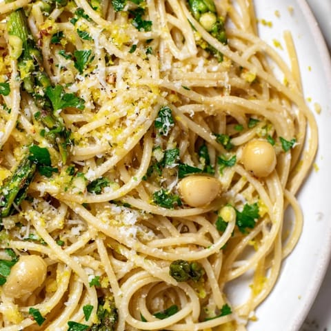 A close-up of Roasted Garlic & Asparagus Pasta, featuring golden roasted garlic cloves and bright green asparagus spears tangled in spaghetti, finished with grated Parmesan and fresh parsley.
