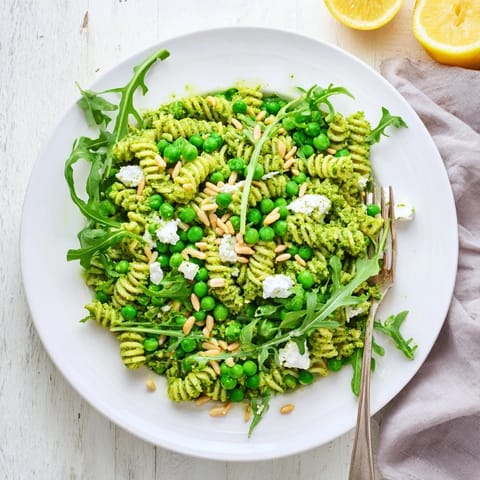 A close-up of vibrant Spring Green Pesto Pasta Salad in a white bowl, showcasing glistening fusilli coated in basil pesto, sweet peas, peppery arugula, and golden toasted pine nuts.