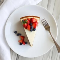 A thick slice of New York Cheesecake on a white plate with a fork, garnished with fresh raspberry sauce drizzled over the top.